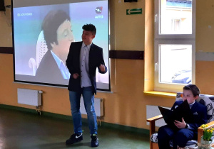 Chłopak ubrany w białą koszulę i marynarkę śpiewa piosenkę. Za nim wyświetlany jest fragment oryginalnego wykonania.