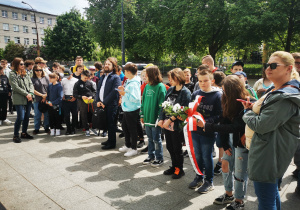 Uczniowie zgromadzeni przed pomnikiem ofiar Holocaustu słuchają w skupieniu przewodnika wycieczki.