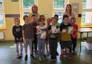 Dzieci z klasy pierwszej wraz z nauczycielką i panią dyrektor pozują do zdjęcia, prezentując dyplomy otrzymane w konkursie na kapliczkę dla Matki Boskiej.