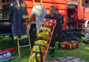 Strażacy prezentują sposób przenoszenia osoby poszkodowanej na noszach.