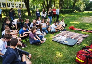 Uczniowie siedzą na trawie, przed nimi, na kocu leży fantom do ćwiczeń masażu serca.