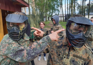 Uczniowie w strojach moro oraz maskach ochronnych na głowie do gry w paintballa pokazują farbę na swoich kombinezonach.