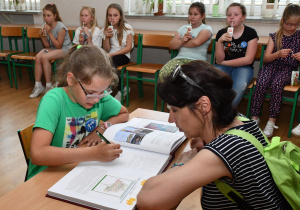 Uczennica w asyście wychowawczyni dokonuje wpisu do księgi pamiątkowej. W tle uczniowie siedzą na krzesłach.