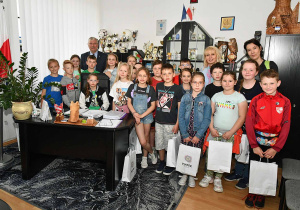 Uczniowie pozują do zdjęcia pamiątkowego w gabinecie Pana Burmistrza. Wszyscy trzymają papierowe torebki z otrzymanymi upominkami. W tle widać regał z licznymi pucharami.