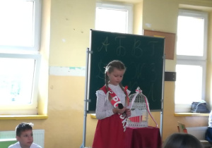 Uczennica ubrana w barwy narodowe trzyma białą klatkę z gołębiem.