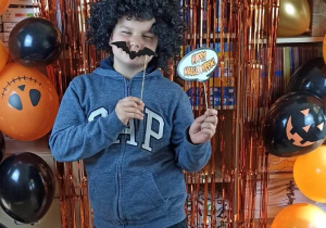 Chłopiec w kapeluszu czarownicy i wąsami pozuje na tle dekoracji Halloweenowej.
