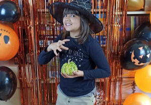 Uczennica w kapeluszu czarownicy i dynią w dłoni pozuje na tle napisu Happy Halloween.