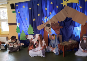Na tle bożonarodzeniowej dekoracji siedzi pastuszek przy ognisku z gitarą. Obok siedzi anioł i śpiewa kolędę.