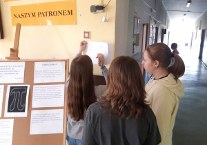 Uczniowie rozwiązują łamigłówki matematyczne przyczepione na ścianie korytarza szkolnego.