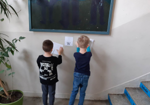 Uczniowie rozwiązują łamigłówki matematyczne przyczepione na ścianie korytarza szkolnego.
