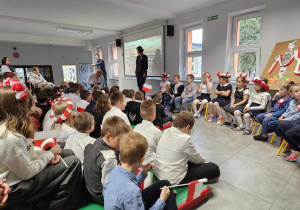Uczniowie ubrani w barwy biało-czerwone siedzą na szkolnym holu, słuchając przemówienia pani dyrektor z okazji Święta Niepodległości.
