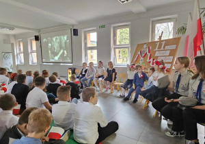 Uczniowie wpatrują się w ekran, na którym wyświetlany jest tekst pieśni patriotycznej. Uczniowie ubrani są odświętnie, trzymają chorągiewki biało-czerwone; widać także uczniów w strojach harcerzy.