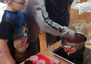 Uczniowie gromadzą się przy garnku, w którym podgrzewa się masa mydlana. Na stoliku obok leżą foremki przygotowane do przygotowania mydełka,