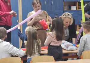 Dzieci gromadzą się przy stole zapełnionym balonami. Jedna z nauczycielek wykonuje dla dzieci figurki zwierząt z balonów.