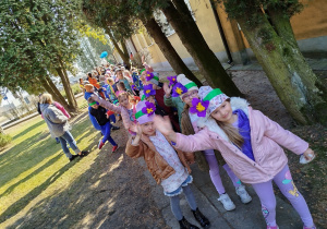 Uczniowie stoją przed budynkiem szkoły trzymając w dłoniach wiosenne kwiaty.