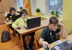 Uczniowie ze słuchawkami na uszach rozwiązują zadania konkursowe na laptopach.