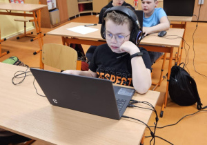 Uczniowie ze słuchawkami na uszach rozwiązują zadania konkursowe na laptopach.
