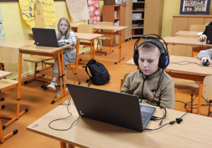 Uczniowie ze słuchawkami na uszach rozwiązują zadania konkursowe na laptopach.