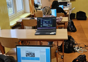 Uczniowie ze słuchawkami na uszach rozwiązują zadania konkursowe na laptopach.