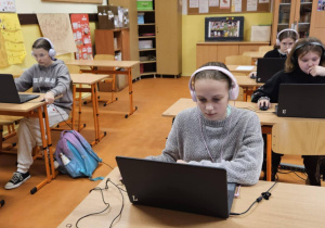 Uczniowie ze słuchawkami na uszach rozwiązują zadania konkursowe na laptopach.
