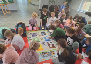 Dzieci gromadzą się przy macie na podłodze, na której widać różne pojemniki na śmieci.