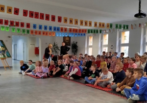 Uczniowie siedzą na szkolnym holu podczas prelekcji na temat segregacji śmieci.