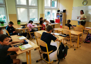 Uczniowie w klasie słuchają w skupieniu nauczycielki czytającej tekst.