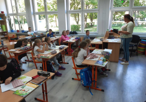 Uczniowie w klasie słuchają w skupieniu nauczycielki czytającej tekst.