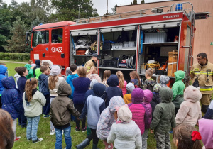 Strażacy prezentują wnętrze wozu strażackiego grupie dzieci na boisku szkolnym.