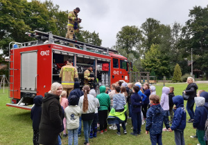 Strażacy prezentują wyposażenie wozu strażackiego grupie dzieci.