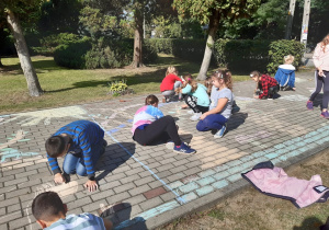Grupa uczniów z klasy trzeciej maluje kolorową kredą kostkę brukową przed budynkiem szkoły.