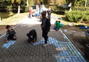 Grupa uczniów z klasy trzeciej maluje kolorową kredą kostkę brukową przed budynkiem szkoły.