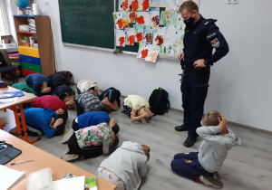 Uczniowie klasy trzeciej skuleni na podłodze ćwiczą bezpieczną pozycję w razie ataku groźnego psa.