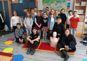 Uczniowie klasy trzeciej wraz z uczniami z klasy siódmej ubranymi w stroje strażackie pozują do zdjęcia po zakończonych zajęciach z udzielania pierwszej pomocy.