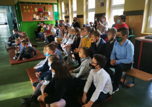 Uczniowie z młodszych klas wraz z nauczycielami siedzą na materacach i ławkach na szkolnym korytarzu przyglądając się przedstawieniu.