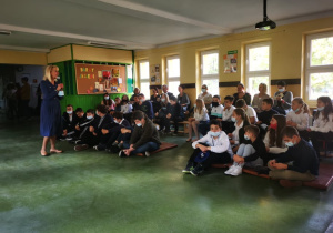 Uczniowie siedzący na materacach i ławkach przysłuchują się wystąpieniu pani dyrektor z okazji Dnia Edukacji Narodowej.