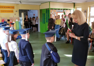 Pani Dyrektor przyjmuje uczniów klasy pierwszej do grona społeczności szkolnej przykładając symbolicznie duży ołówek do ramienia każdego z uczniów. W tle widać zgromadzonych rodziców.