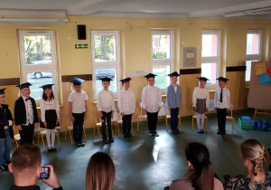 Uczniowie klasy pierwszej stoją przed punlicznością w odświętnych strojach, prezentując przygotowany program artystyczny.
