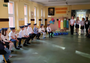 Uczniowie klasy pierwszej siedzą na środku sceny, słuchając przemówienia uczennic z klasy czwartej. W tle widać rodziców robiących zdjęcia.