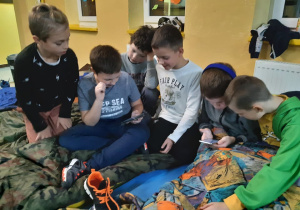 Uczniowie klasy trzeciej z telefonami komórkowymi siedzą na materacach na szkolnym korytarzu podczas nocowania w szkole,