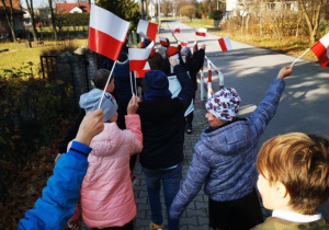 Grupa uczniów maszeruje uroczyście w pochodzie unosząć w górę biało- czerwone chorągiewki.