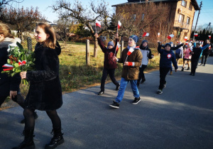 Grupa uczniów maszeruje uroczyście w pochodzie unosząć w górę biało- czerwone chorągiewki.
