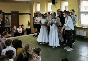 Uczniowie klasy piątej stoją na środku sceny, śpiewając kolędę przed widownią szkolną.