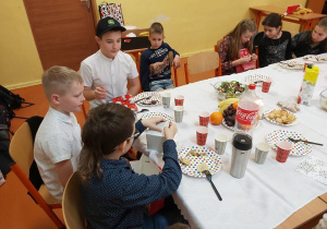 Uczniowie siedzą przy stole wigilijnym w klasie. Oglądają prezenty.