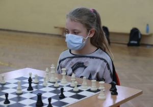 Uczennica przy stole szachowym przygląda się w skupieniu planszy i pionkom.