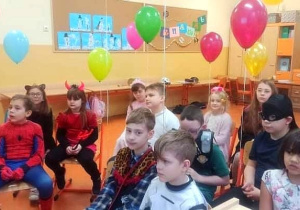 Uczniowie patrzą z zainteresowaniem na film wyświetlany na tablicy interaktywnej. Wszyscy w strojach karnawałowych. Każde dziecko trzyma balon z helem.
