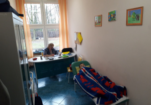 Pielętniarka siedzi przy biurku. Dziecko śpi na leżance.