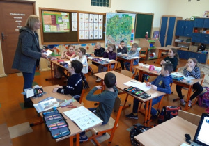 Pielęgniarka stoi przed dziećmi siedzącymi w ławkach.