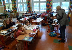 Pielęgniarka stoi przed dziećmi siedzącymi w ławkach.