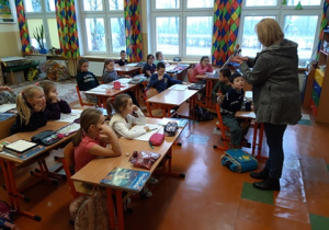 Pielęgniarka stoi przed dziećmi siedzącymi w ławkach.